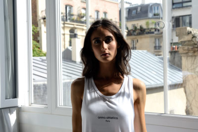 Une mannequin aux longs cheveux noirs se tient debout à l'intérieur devant de grandes fenêtres, laissant la lumière naturelle l'illuminer. Elle porte un haut blanc sans manches avec le texte « anima athletica Paris » dessus. Capture par le célèbre photographe Thomas Duclert, on aperçoit derrière elle une vue des bâtiments voisins.