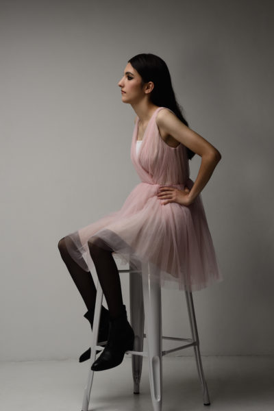 Une mannequin aux cheveux longs et foncés, vêtue d'une robe rose clair sans manches et de bottines noires, est assise sur un grand tabouret blanc sur un fond gris uni, regardant vers la droite avec les mains sur les hanches, capturée impeccablement par l'objectif du photographe Thomas Duclert.