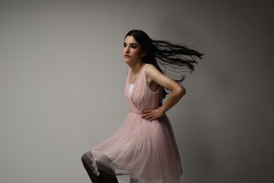 Une mannequin aux longs cheveux noirs se tient debout sur un fond gris uni. Elle porte une robe rose clair sans manches avec une jupe fluide et semble être en mouvement, ses cheveux traînant derrière elle, suggérant le mouvement. Son expression sérieuse évoque la mode parisienne telle que l'a capturée Thomas Duclert.