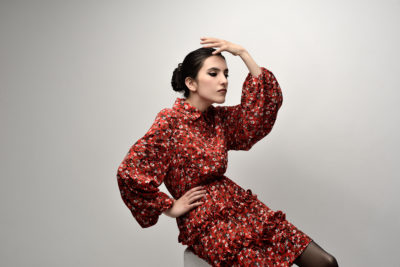 Une mannequin aux cheveux noirs relevés en chignon, vêtue d'une robe fleurie rouge à manches longues bouffantes, pose sur un fond gris clair. Dans le style parisien, elle pose une main sur sa hanche et l'autre sur son front, inclinant légèrement la tête sur le côté comme si elle était prise par l'objectif d'un photographe.