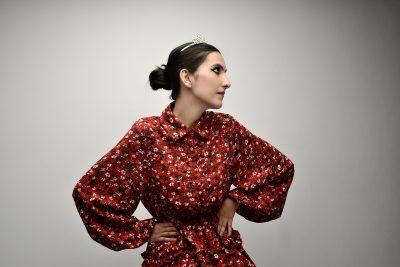 Une mannequin coiffée en chignon et coiffée d'un petit diadème pose dans une robe rouge à motifs floraux. Dans le style parisien, elle se tient de profil, les mains sur les hanches, sur un fond uni et clair, rappelant l'élégance admirée par Thomas Duclert.