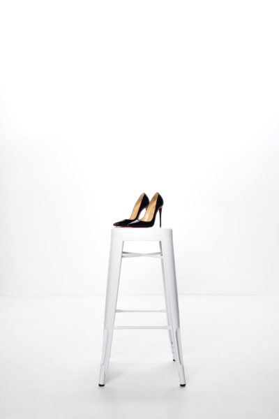 Une paire de chaussures Louboutin noires à talons hauts et semelles rouges repose sur un haut tabouret en métal blanc sur un fond blanc uni. Photographié par Thomas Duclert à Paris, le contraste met en valeur le design élégant des chaussures.