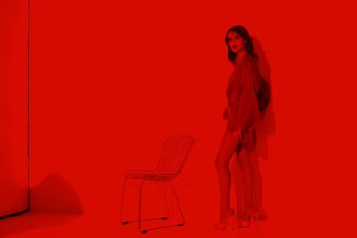 Une mannequin se tient debout sur un fond rouge vif, vêtue d’une tenue rouge chic et de chaussures à talons hauts. Elle se penche légèrement en arrière avec une main sur la hanche, incarnant la mode parisienne. Une chaise à structure métallique est placée près d’elle, projetant des ombres sur le mur à côté de sa silhouette élégante – une scène qui rappelle l’œuvre de Thomas Duclert.