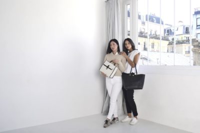 Deux manenquin se tiennent devant une grande fenêtre dans une pièce lumineuse. L'une porte un haut crème et un pantalon blanc, tenant un sac à main beige avec un ruban noir. L'autre porte un haut blanc et un pantalon noir, tenant un sac à main noir. Prises en photo par le photographe parisien Thomas Duclert, elles regardent vers l'objectif.