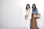 Deux mannequins se tiennent côte à côte sur un fond blanc. La mannequin de gauche, affichant un style chic, porte un blazer et un pantalon blancs, portant un petit sac noir et blanc. La mannequin de droite porte une chemise en jean bleu avec une longue jupe marron, tenant un grand sac à main en tissu.