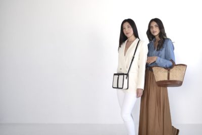 Deux mannequins se tiennent côte à côte sur un fond blanc. La mannequin de gauche, affichant un style chic, porte un blazer et un pantalon blancs, portant un petit sac noir et blanc. La mannequin de droite porte une chemise en jean bleu avec une longue jupe marron, tenant un grand sac à main en tissu.