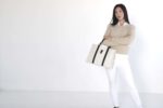 Une mannequin aux longs cheveux noirs se tient debout avec assurance sur un fond clair et uni. Elle porte un pull beige sur une chemise blanche, un pantalon blanc et tient un élégant sac à main blanc à bretelles noires. Photographiée en mode intemporel, elle regarde directement l'objectif, incarnant l'essence chic de Paris.