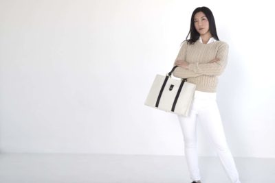 Une mannequin aux longs cheveux noirs se tient debout avec assurance sur un fond clair et uni. Elle porte un pull beige sur une chemise blanche, un pantalon blanc et tient un élégant sac à main blanc à bretelles noires. Photographiée en mode intemporel, elle regarde directement l'objectif, incarnant l'essence chic de Paris.