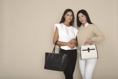 Deux mannequins se tiennent debout sur un fond beige, toutes deux tournées vers le photographe. Celle de gauche porte un haut sans manches blanc et un pantalon noir, tenant un sac à main noir. Celle de droite porte un pull beige et un pantalon blanc, tenant un sac à main blanc, incarnant la mode chic parisienne.
