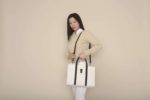 Une mannequin aux cheveux longs se tient debout sur un fond beige, à la manière parisienne, vêtue d'un pull marron clair sur une chemise blanche à col et un pantalon blanc. Elle tient un grand sac fourre-tout blanc avec des poignées noires et une poche avant, comme si elle posait pour un photographe.