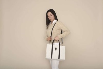 Une mannequin aux cheveux longs se tient debout sur un fond beige, à la manière parisienne, vêtue d'un pull marron clair sur une chemise blanche à col et un pantalon blanc. Elle tient un grand sac fourre-tout blanc avec des poignées noires et une poche avant, comme si elle posait pour un photographe.