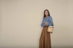 Une mannequin aux cheveux longs et noirs, vêtue d'une chemise en chambray bleu rentrée dans une jupe ample marron à taille haute, se tient debout sur un fond beige uni. Elle tient un sac à main tressé de style seau. Elle regarde au loin, dégageant une mode parisienne et une confiance calme qui rappelle le style de Thomas Duclert.