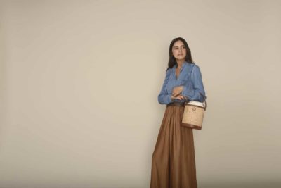 Une mannequin aux cheveux longs et noirs, vêtue d'une chemise en chambray bleu rentrée dans une jupe ample marron à taille haute, se tient debout sur un fond beige uni. Elle tient un sac à main tressé de style seau. Elle regarde au loin, dégageant une mode parisienne et une confiance calme qui rappelle le style de Thomas Duclert.