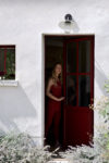 Une mannequin se tient à l'intérieur et à l'extérieur, dans l'embrasure d'une maison aux murs blancs et à la porte rouge. Vêtue d'une tenue rouge inspirée de la mode, elle regarde au loin. La lumière du soleil et les plantes sont visibles à l'extérieur, tandis qu'un luminaire orne l'entrée, évoquant un air d'élégance parisienne rappelant les œuvres de Thomas Duclert.