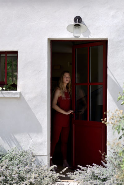 Une mannequin se tient à l'intérieur et à l'extérieur, dans l'embrasure d'une maison aux murs blancs et à la porte rouge. Vêtue d'une tenue rouge inspirée de la mode, elle regarde au loin. La lumière du soleil et les plantes sont visibles à l'extérieur, tandis qu'un luminaire orne l'entrée, évoquant un air d'élégance parisienne rappelant les œuvres de Thomas Duclert.