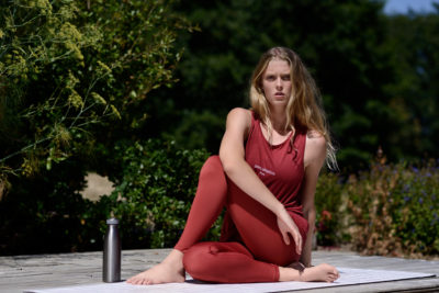 Une mannequin aux cheveux longs est assise en tailleur sur un tapis à l'extérieur, vêtue d'un haut rouge sans manches et d'un legging assorti. Elle est entourée de verdure, avec une bouteille d'eau métallique à côté d'elle. Adoptant un mode tranquille, elle semble méditer ou prendre une pose détendue.