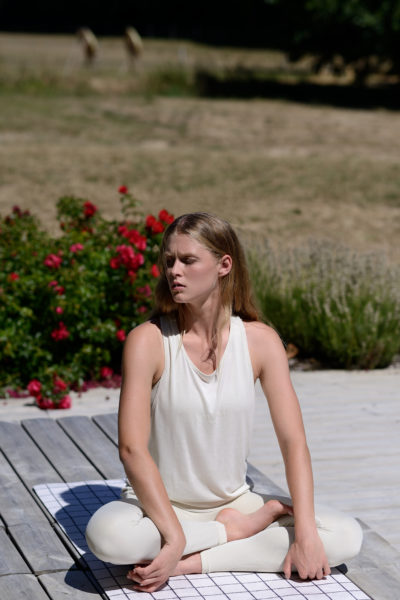 Une mannequin aux longs cheveux blonds est assise en tailleur sur un tapis, pratiquant le yoga dans un cadre extérieur serein. Elle porte un haut blanc sans manches et des leggings blancs. En arrière-plan, des fleurs rouges et de la verdure s'épanouissent. Le temps semble ensoleillé, créant une scène idyllique parfaite pour tout photographe de mode parisien.