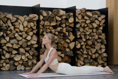 Une mannequin en tenue de sport blanche pratique le yoga à l'intérieur, en effectuant la posture du chien tête en haut sur un tapis rayé. Derrière elle, des piles de bois de chauffage soigneusement empilées sur des étagères noires ajoutent au style épuré et minimaliste du décor, rappelant un studio parisien serein capturé par un photographe expérimenté.