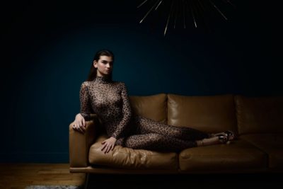 Une mannequin vêtue d'une combinaison à imprimé léopard se prélasse sur un canapé en cuir beige, devant un mur bleu foncé. La pièce, photographiée par le photographe Thomas Duclert, est dotée d'un éclairage minimal, projetant des ombres et soulignant l'esthétique épurée et moderne. Un élément décoratif avec de longs rayons rayonnants est fixé au mur au-dessus.