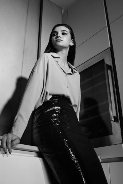 Une mannequin en tenue élégante se tient debout, confiante, dans une cuisine moderne. Elle porte un chemisier clair rentré dans une jupe brillante et sombre à paillettes. La photo, prise par le photographe Thomas Duclert, est en noir et blanc, mettant en valeur les contrastes de textures et d'ombres.