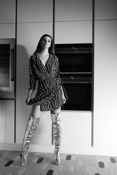 Une mannequin se tient debout dans une cuisine moderne, vêtue d'une robe moulante à motifs et de bottes brillantes jusqu'aux genoux. L'image en noir et blanc capture sa tenue frappante et sa pose confiante, rappelant la mode parisienne. Elle a une main sur sa robe, avec des armoires élégantes et des appareils intégrés encadrant la scène.