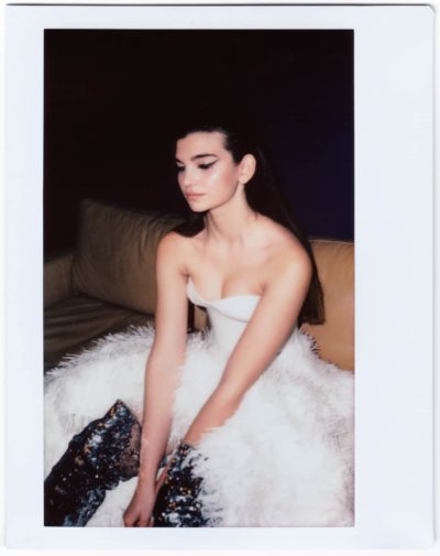 Sur cette photographie maussade de Thomas Duclert, une manequin aux cheveux longs et noirs est assise sur un canapé marron. Elle porte une robe bustier blanche avec une jupe à plumes et des bottes à paillettes scintillantes, le regard légèrement baissé sur un fond sombre – un moment évocateur capturé à Paris.