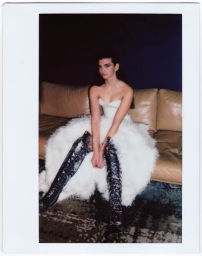 Une mannequin est assise sur un canapé beige, vêtue d'une robe blanche bustier avec une jupe bouffante et des bottes scintillantes jusqu'aux genoux, évoquant la mode de la haute couture. Photographiée par Thomas Duclert, elle pose ses mains sur ses genoux, regardant légèrement sur le côté sur un fond sombre.