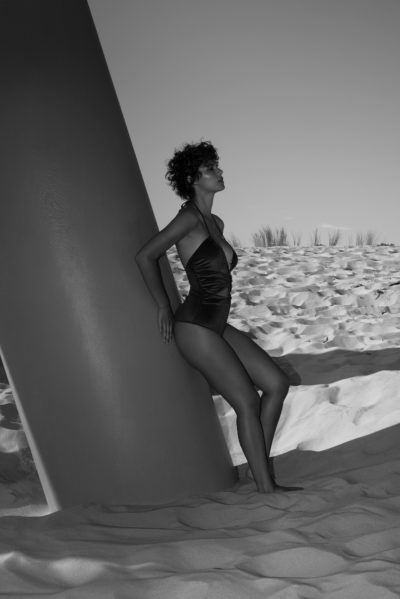 Une mannequin en maillot de bain est adossée à une grande structure cylindrique sur une plage de sable. Prise en photo par le photographe Thomas Duclert, elle regarde au loin, les mains posées derrière elle. En arrière-plan, les dunes de sable de Paris sont ornées d'une végétation clairsemée. L'image est en noir et blanc.