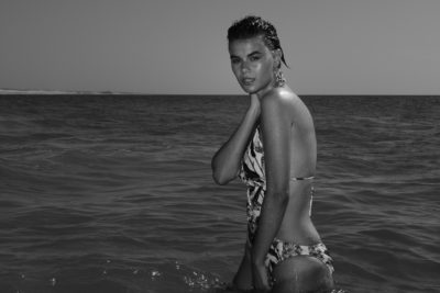 Image monochrome d'une mannequin debout dans l'océan, l'eau jusqu'à la taille, portant un bikini à motifs. Elle incline légèrement la tête et pose une main sur son cou, tout en regardant directement l'objectif. Capturée avec finesse par le photographe Thomas Duclert, l'arrière-plan montre un ciel clair et un rivage lointain.