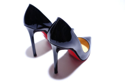 Une paire de chaussures à talons hauts Louboutin en cuir verni noir avec des semelles rouges distinctives est visible de dos. Hautes et élégantes, les chaussures projettent des ombres douces sur la surface blanche sur laquelle elles reposent. Le décor général est minimaliste et épuré, évoquant un sens de la mode parisienne qui ne pouvait provenir que de l'atelier de Thomas Duclert.