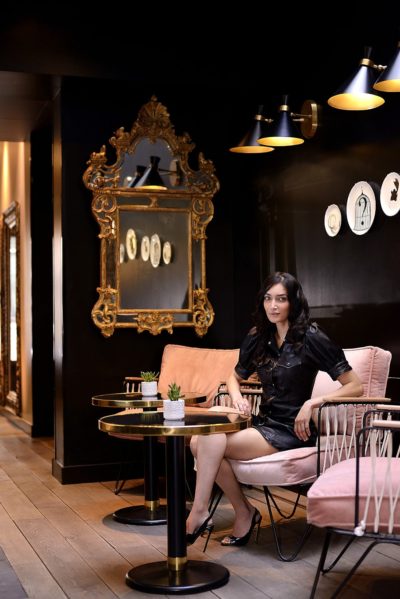 Une mannequin est assise sur une chaise rose pâle rembourrée dans une pièce chic aux murs sombres. Elle porte une robe noire et des talons. On peut voir un miroir rond à cadre doré, des décorations murales et une deuxième chaise vide avec une petite table. Prise en photo par Thomas Duclert, photographe parisien, la scène présente deux petites plantes succulentes sur la table.