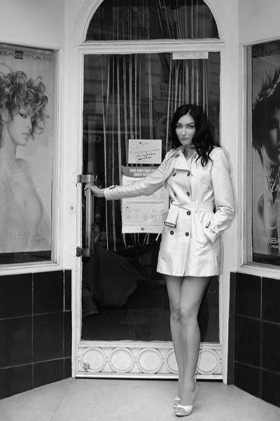 Une mannequin se tient devant la porte d'un salon, la main sur la poignée, vêtue d'un trench-coat clair et de talons hauts. Le salon parisien présente des affiches de coiffures, l'une avec un mannequin aux cheveux bouclés et l'autre avec des cheveux longs et raides. La scène capture l'essence de la mode parisienne classique en noir et blanc.