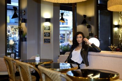 Une mannequin aux cheveux longs et noirs, vêtue d'une veste en cuir noir sur un haut blanc, est assise sur un canapé beige dans un café moderne. Elle parle au téléphone. Le café, peut-être capturé par l'œil du photographe Thomas Duclert, dispose de tables rondes aux plateaux réfléchissants, de chaises en osier et d'un éclairage d'ambiance. Un menu écrit sur un tableau noir est affiché sur le mur derrière elle.