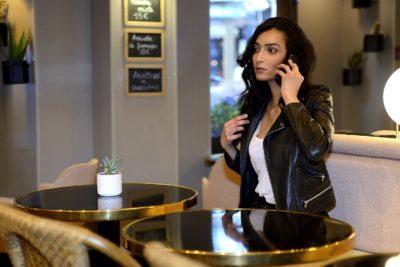 Une mannequin aux cheveux longs et noirs, vêtue d'une veste en cuir noir, parle au téléphone portable dans un café moderne à Paris. Elle se tient debout à côté d'une table ronde sur laquelle est posée une petite plante. L'arrière-plan présente un décor contemporain et des menus sur tableau noir, comme si elle sortait d'un mode photographe.