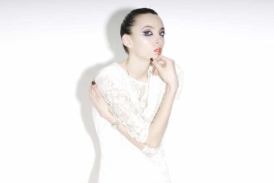 Une mannequin aux cheveux lissés en arrière pose devant un fond blanc. Elle porte une robe en dentelle blanche et sa main droite touche légèrement ses lèvres tandis que son bras gauche croise son torse. Capturée à la manière parisienne par un photographe talentueux, elle regarde vers l'objectif avec une expression neutre.