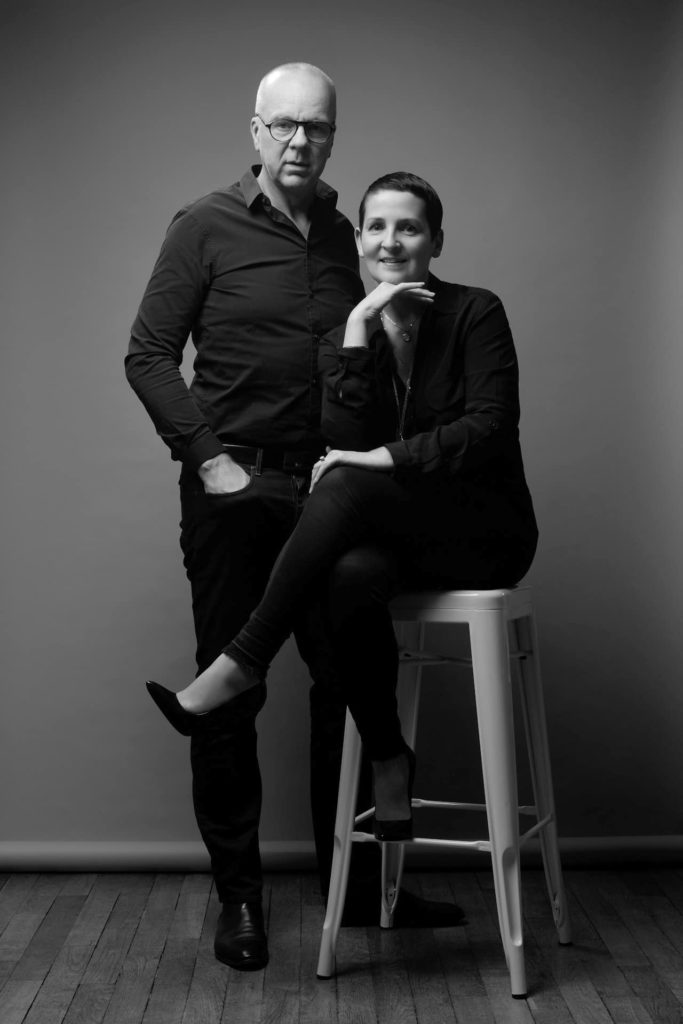 Photographie en noir et blanc d'un homme et de Claire et Thomas Duclert posant devant un fond uni. Thomas debout, portant des lunettes, la main nonchalamment dans sa poche. Claire est assise sur un haut tabouret, les jambes croisées, le menton posé sur sa main, tous deux vêtus de vêtements sombres.