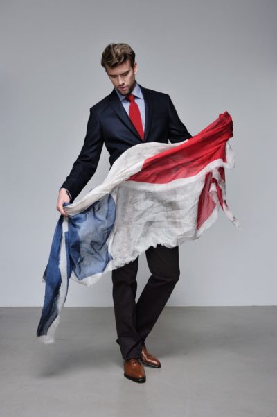 Un mannequin en costume et cravate rouge, rappelant la mode parisienne classique, tient un grand tissu patiné à rayures bleues, blanches et rouges. Il se tient debout devant un fond gris uni, regardant le tissu qui tombe sur ses bras et touche le sol.