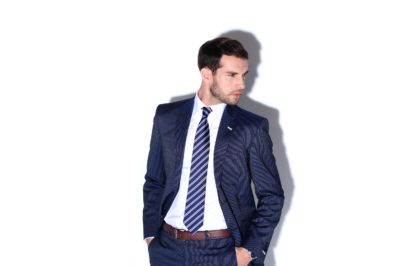 Un mannequin aux cheveux courts et noirs, vêtu d'un costume bleu foncé, d'une chemise blanche et d'une cravate rayée, se tient debout, les mains dans les poches. Il regarde vers sa droite, avec une expression sérieuse. L'arrière-plan est blanc uni, projetant une ombre derrière lui ; une image capturant le chic subtil de la mode parisienne.