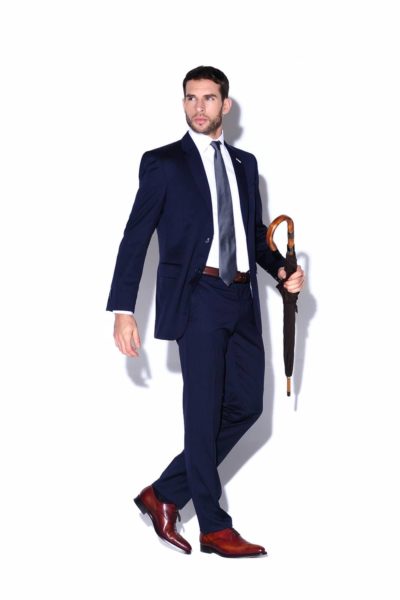 Un mannequin en costume bleu foncé, chemise blanche et chaussures marron, se tient debout sur un fond blanc. Il tient dans sa main droite un parapluie fermé avec un manche en bois et regarde vers sa gauche. La scène est capturée avec la précision d'un photographe parisien chevronné.