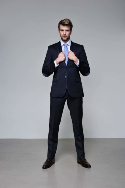 Un mannequin se tient debout, confiant, dans un costume sombre avec une bordure bleue contrastante, ajustant sa cravate bleu clair. Il porte des chaussures noires et pose sur un fond gris clair uni, illustrant un look moderne et élégant, parfaitement capturé par un photographe parisien.