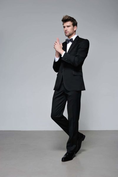 Un mannequin en costume noir et nœud papillon se tient avec assurance sur un fond gris uni, arborant le look classique d'un espion. Avec ses cheveux coiffés et sa pose comme s'il tenait une arme à feu, il respire la sophistication. Cette photographie capture l'essence de la mode parisienne avec une jambe légèrement pliée et une élégance posée.