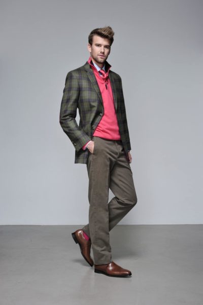 Un mannequin pose devant un fond gris uni, vêtu d'un blazer à carreaux verts sur une chemise rouge et un pantalon marron. Il a une main dans sa poche et se tient debout avec une jambe légèrement pliée, mettant en valeur des chaussures marron. Ses cheveux sont coiffés avec soin, une touche parisienne moderne capturée par le photographe.