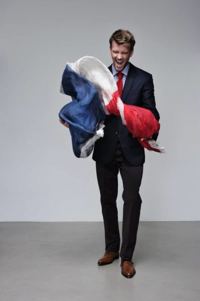Un mannequin en tenue de soirée, comprenant un costume sombre, une chemise blanche et une cravate rouge, secoue et tient avec enthousiasme un grand morceau de tissu tricolore (bleu, blanc et rouge) rappelant Paris. L'expression du visage énergique et passionnée du mannequin reflète la ferveur de la mode.