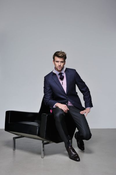 Un mannequin porte un blazer bleu marine sur une chemise rose avec une cravate foncée alors qu'il est assis nonchalamment sur l'accoudoir d'une chaise noire moderne. Il porte un pantalon foncé et des chaussures marron cirées. L'arrière-plan est un décor de studio simple et clair, rappelant la toile d'un photographe.