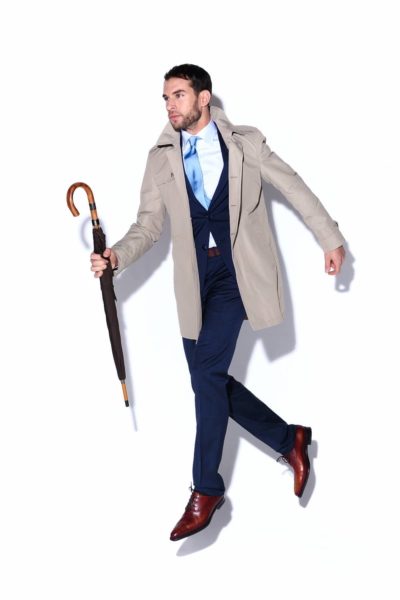 Un mannequin portant un costume bleu sur mesure, une cravate bleu clair et un trench beige se tient debout sur un fond blanc. Il tient un parapluie fermé avec un manche en bois dans sa main droite et semble flotter ou sauter. Il incarne la mode parisienne avec ses chaussures de ville marron.