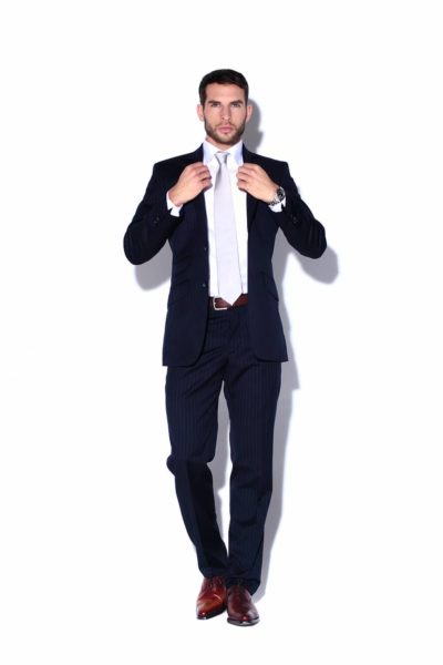 Un mannequin bien habillé, vêtu d'un costume à rayures bleu marine, se tient debout sur un fond blanc, ajustant sa cravate blanche des deux mains. L'homme porte une chemise blanche sous sa veste de costume, des chaussures en cuir marron et une ceinture marron. Il a les cheveux courts et une barbe.