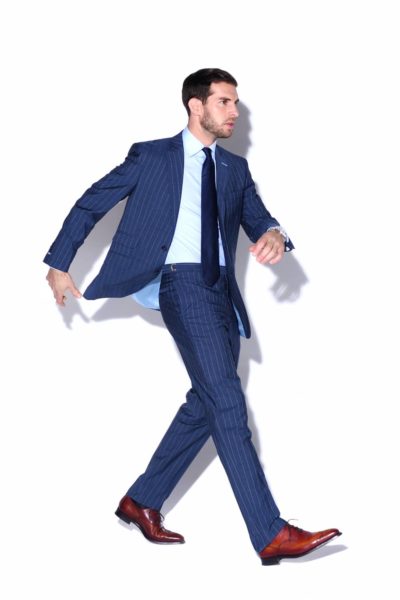 Un mannequin en costume bleu à rayures, chemise bleu clair et cravate bleu marine marche avec assurance sur fond blanc. Il porte des chaussures marron et porte une courte barbe, incarnant la mode moderne comme s'il avait été capturé par le photographe Thomas Duclert.