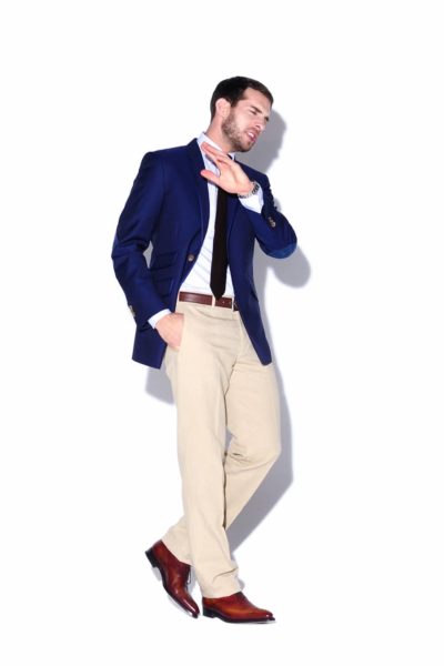 Un mannequin habillé avec élégance se tient debout sur un fond blanc, légèrement penché en arrière, la main droite près du visage comme s'il repoussait quelque chose. Il porte un blazer bleu, une chemise blanche, une cravate noire, un pantalon beige et des chaussures marron, une véritable incarnation de la mode parisienne parfaitement capturée par le photographe.