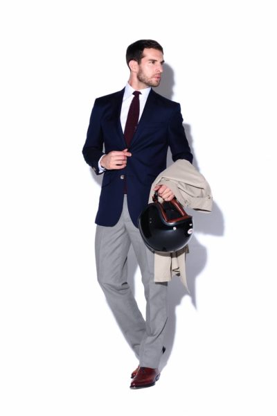 Un mannequin vêtu d'un blazer bleu marine, d'une chemise blanche, d'une cravate bordeaux et d'un pantalon gris, porte un trench-coat beige et un casque de moto noir. Avec ses cheveux noirs courts et son arrière-plan blanc uni, il respire la confiance. Capturé par un photographe talentueux à Paris, son attitude posée attire l'attention.
