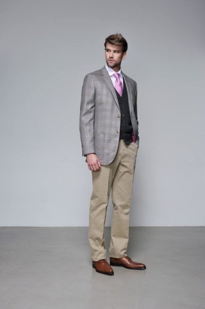 Un mannequin se tient debout sur un fond uni, vêtu d'un blazer à carreaux gris sur un pull noir, d'une chemise rose avec une cravate rose assortie, d'un pantalon beige et de chaussures marron. Il a les cheveux châtain clair et est dans une pose décontractée, regardant légèrement sur le côté. Son ensemble reflète le goût raffiné de la mode de Thomas Duclert.